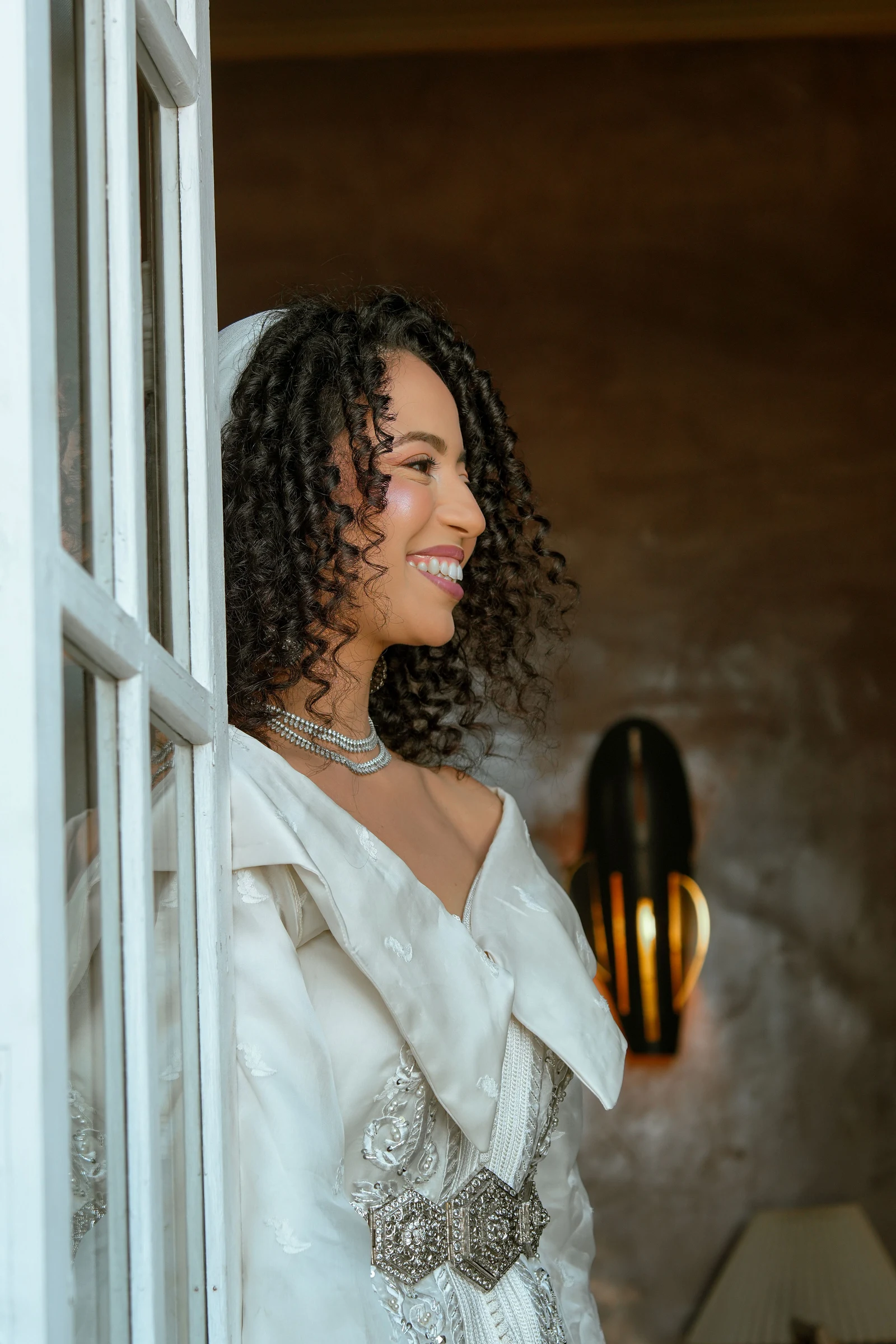 Femme en caftan blanc souriant près d’une fenêtre